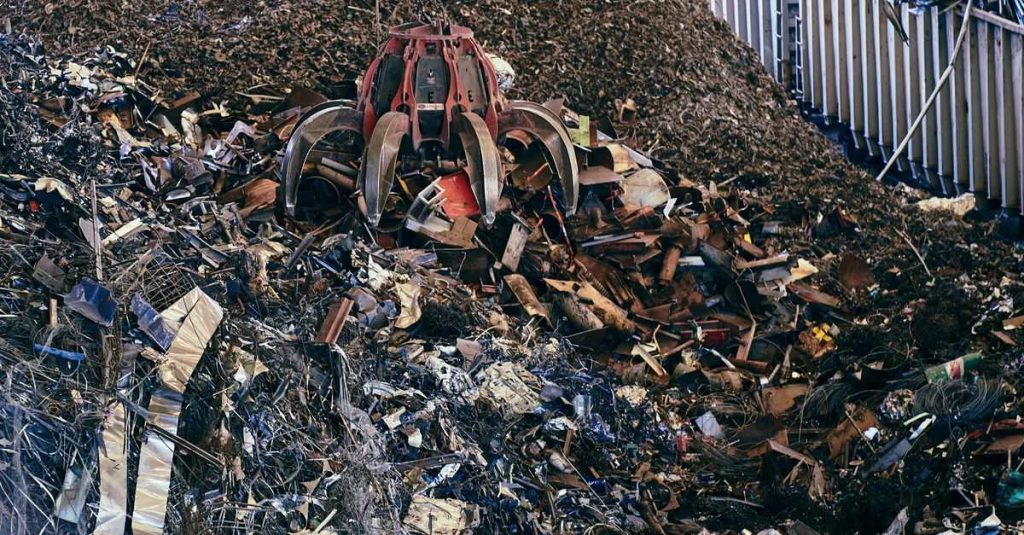 A claw excavator sifting through a large pile of industrial scrap metal outdoors.