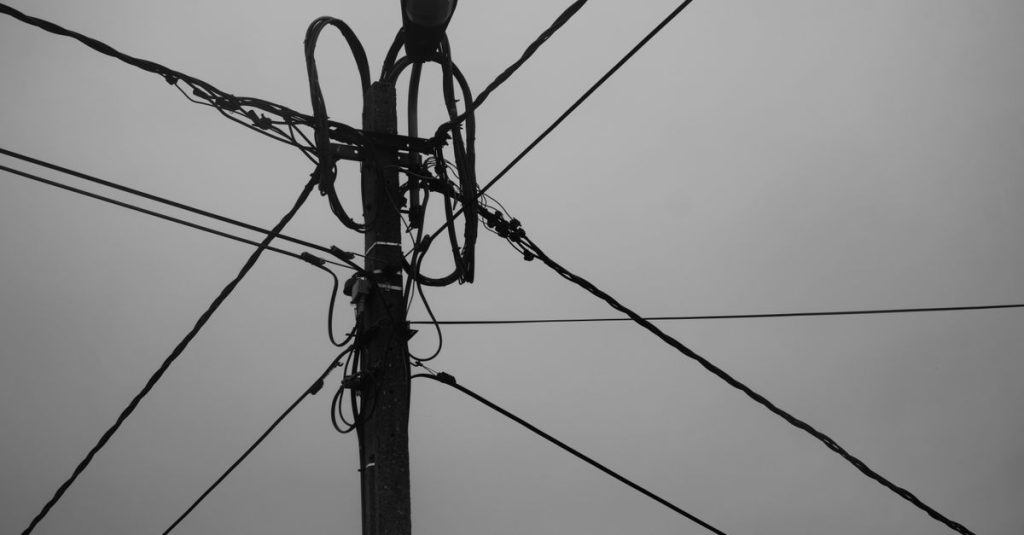 Black and white photo of a utility pole with intersecting power lines and wires.