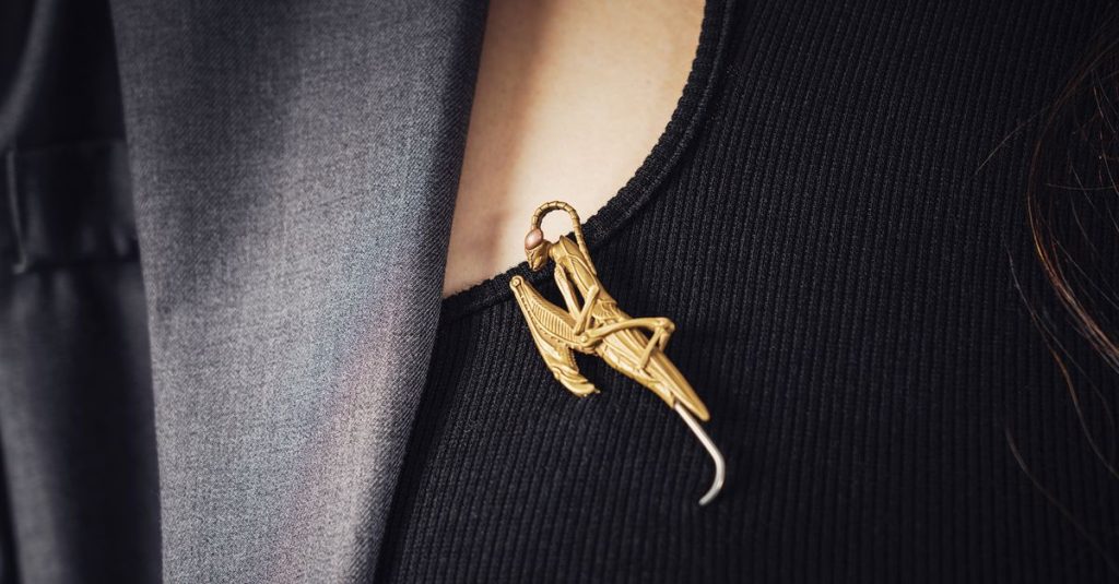 Close-up of a stylish gold insect brooch pinned on a black ribbed blouse, under a grey blazer.
