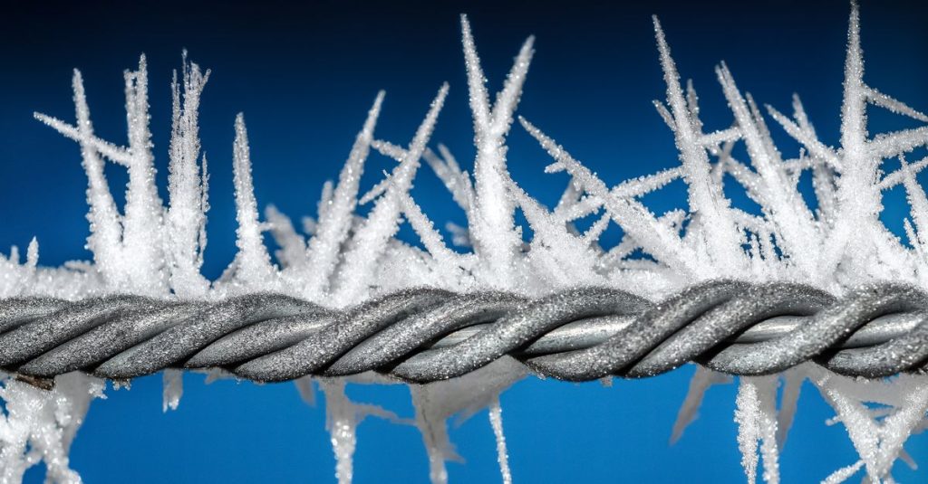 Macro shot of frost-covered metal wire with ice crystals against a blue sky.