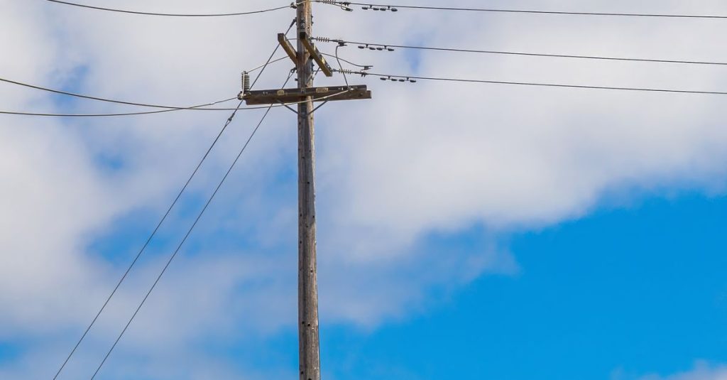 Wooden power pole with transmission lines against a clear blue sky and clouds.