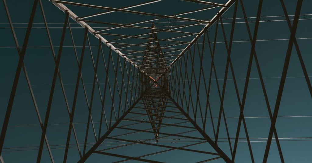 Abstract view looking up inside a transmission tower under a clear blue sky, creating a symmetrical pattern.