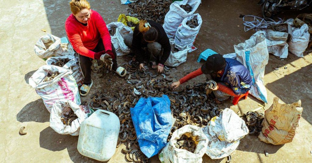 A diverse group sorts through scrap metal in an outdoor market setting, surrounded by bags of recyclables.