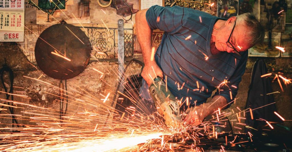 An artisan wearing glasses works with an angle grinder, creating sparks in a rustic workshop.