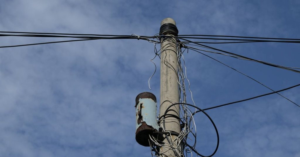Photograph of a utility pole and power lines under a bright blue sky with scattered clouds.