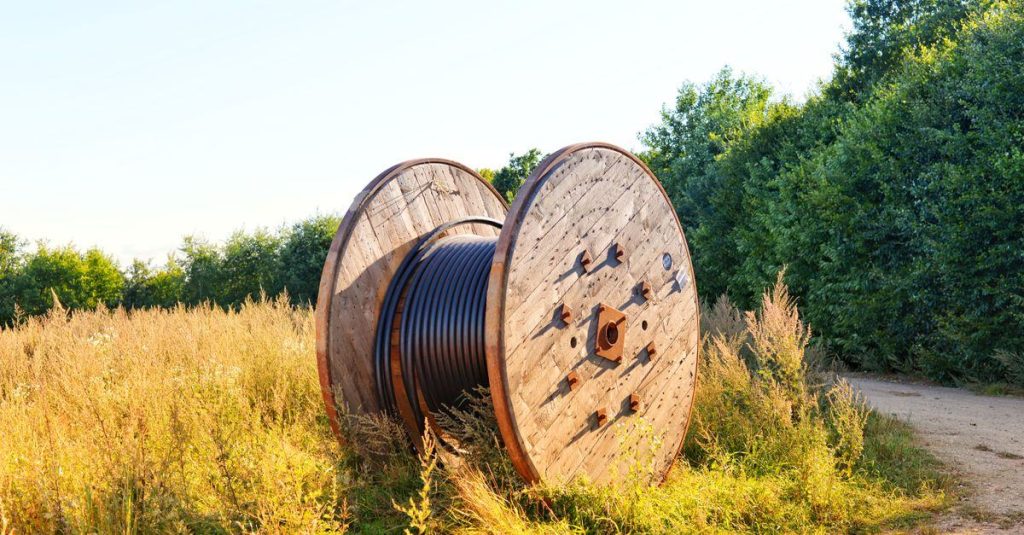 Wooden cable reel with coiled wires in a sunlit field. Ideal infrastructure stock photo.