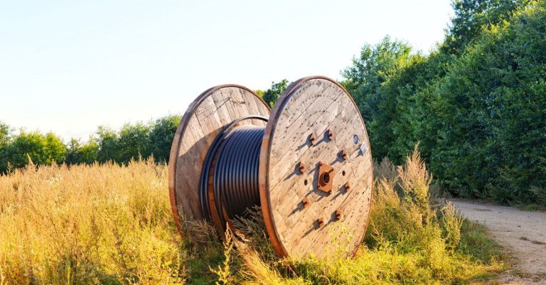 Wooden cable reel with coiled wires in a sunlit field. Ideal infrastructure stock photo.