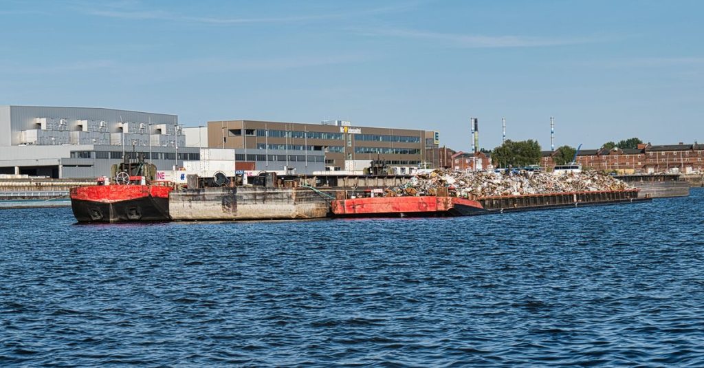 Wide shot of red industrial barges laden with scrap metal on a river near urban industrial buildings.
